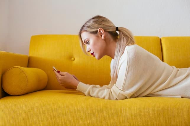 a young blonde woman lying on a yellow coach is using her phone